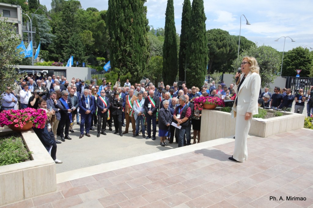 Un evento all'aperto con un pubblico numeroso, dove una donna in abito bianco sta parlando a un microfono, circondata da fiori e alberi.