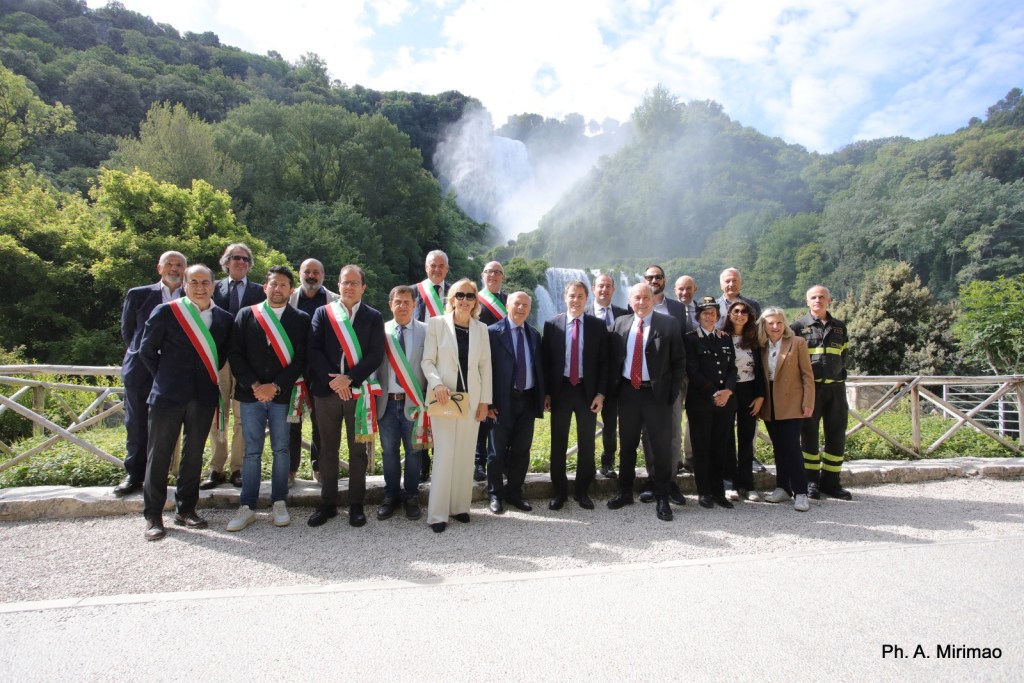 Gruppo di persone in posa davanti a una cascataimmersa nella natura verde, con diversi individui a indossare fasce tricolori.