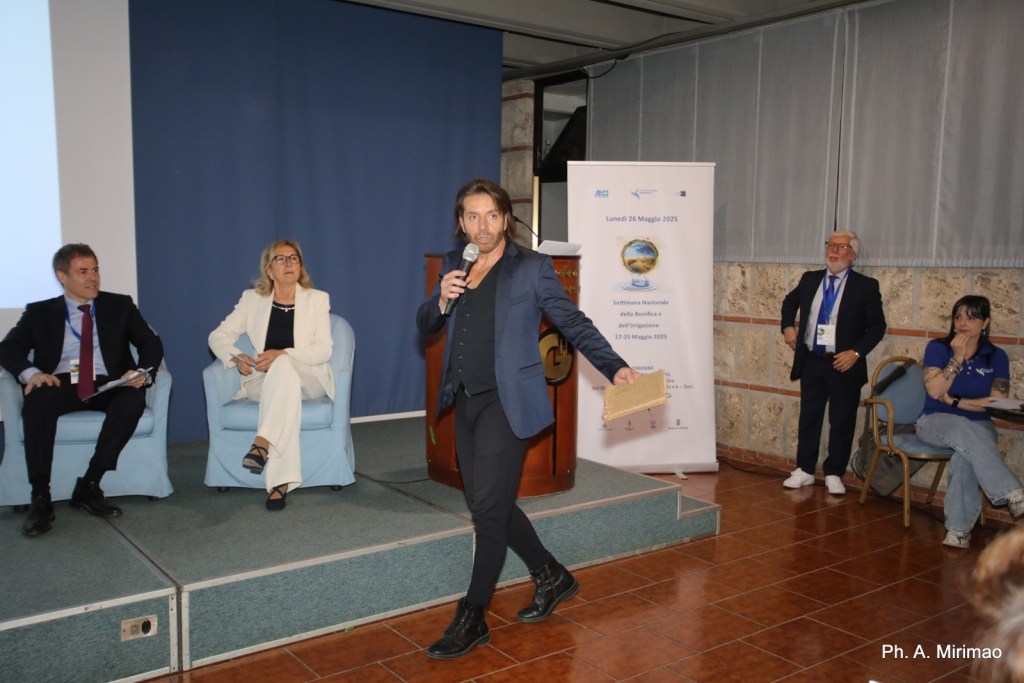 Un partecipante al convegno parla al pubblico mentre altri ospiti ascoltano in un ambiente conferenziale.