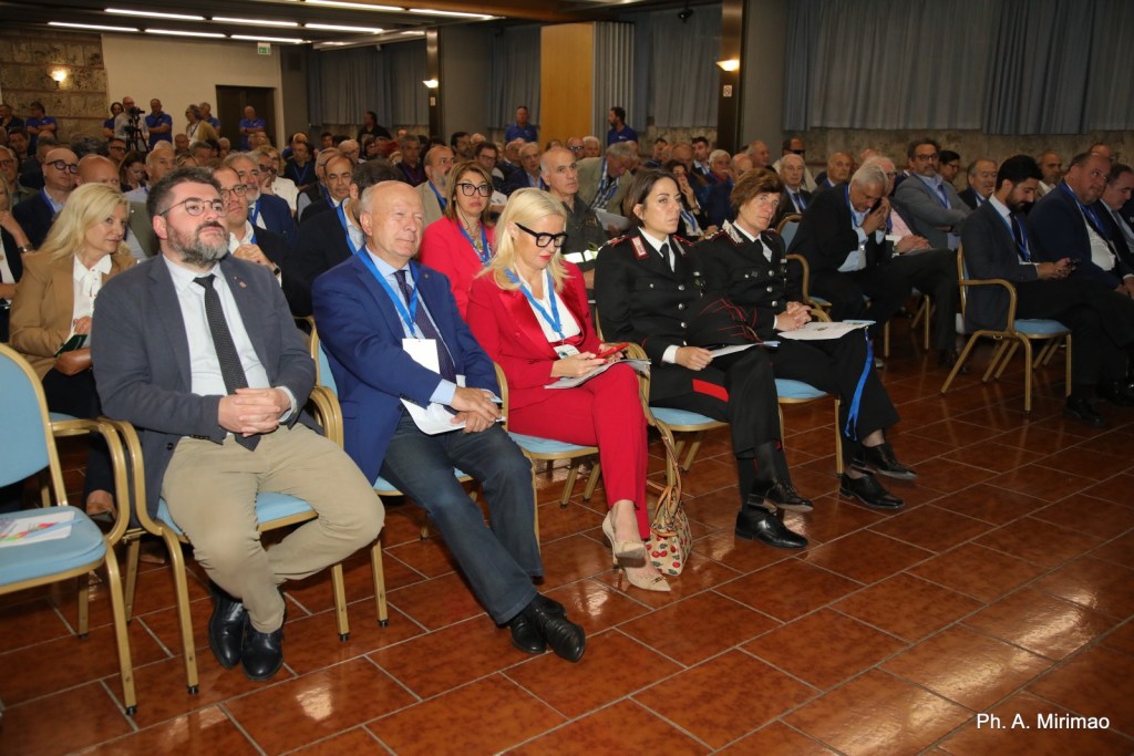 Un gruppo di persone sedute in un convegno, con partecipanti che indossano distintivi e abbigliamento formale, tra cui anche una donna in uniforme di carabinieri.