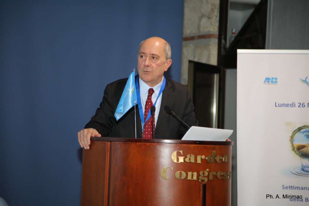 Un relatore al convegno sul tema dell'acqua e della sostenibilità, con un microfono davanti e un foglio di appunti, mentre una bandiera è appesa al suo lato. Sullo sfondo, un pannello informativo del convegno.