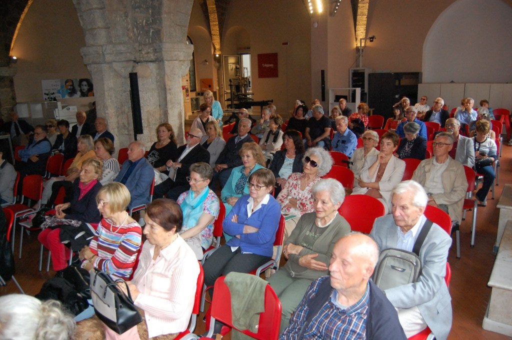 Audience seated in a lecture hall during a university event, with a focus on older adults. The room features stone walls and red chairs.