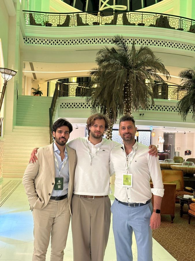 Tre uomini sorridenti si trovano in un ambiente elegante, circondati da palme e decorazioni moderne. Indossano abbigliamento formale e hanno distintivi attaccati ai vestiti, suggerendo la loro partecipazione a un evento importante.