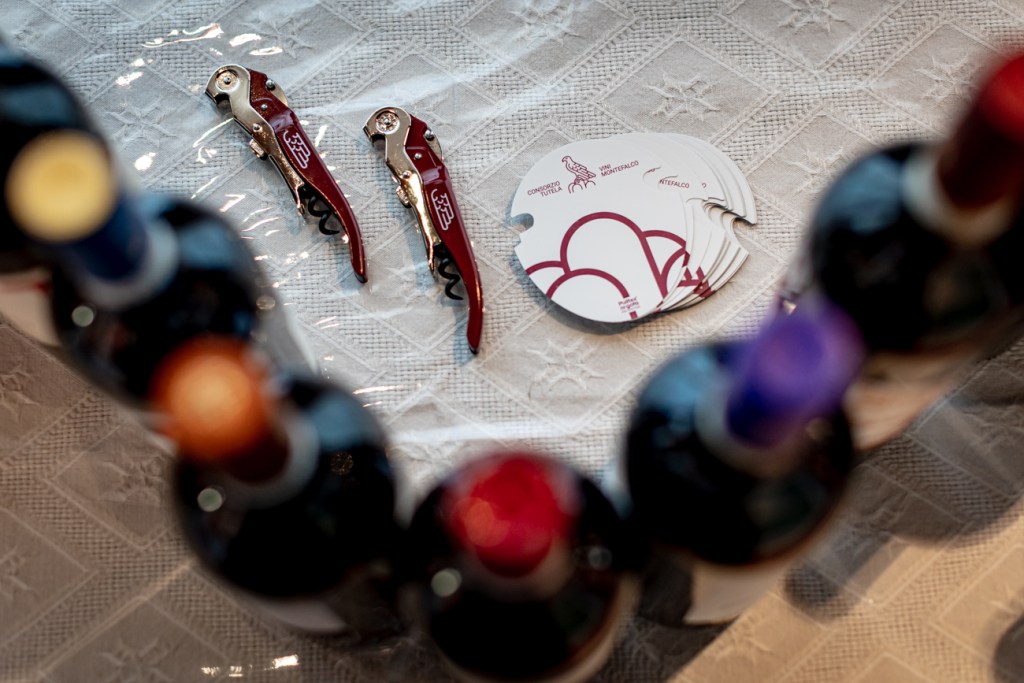 Due cavatappi rossi e schede informative su un tavolo con bottiglie di vino in primo piano.