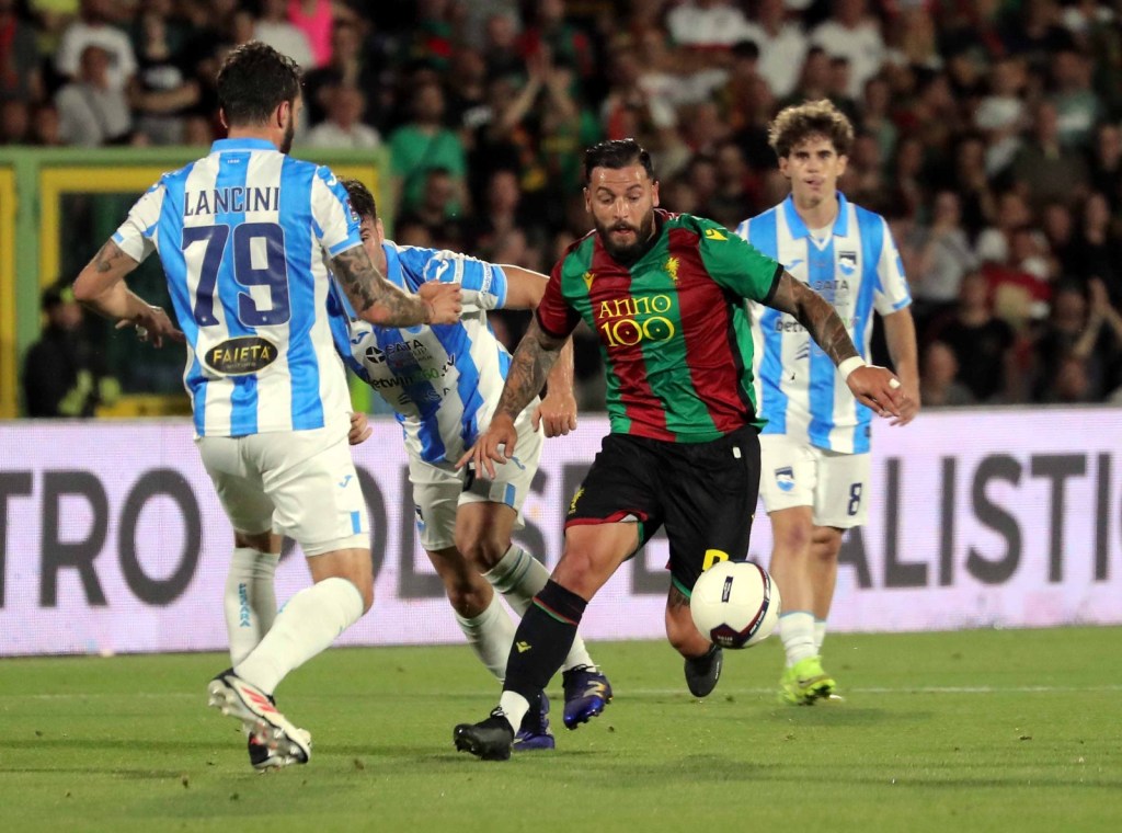 A football player in a red and green jersey dribbles the ball while being chased by two defenders in blue and white jerseys during a match.