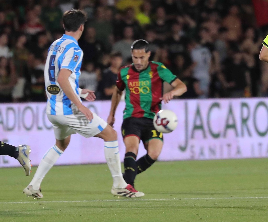 A soccer match in progress, featuring two players: one in a white jersey with blue stripes and another in a green and red striped jersey, both focused on the ball on the field.