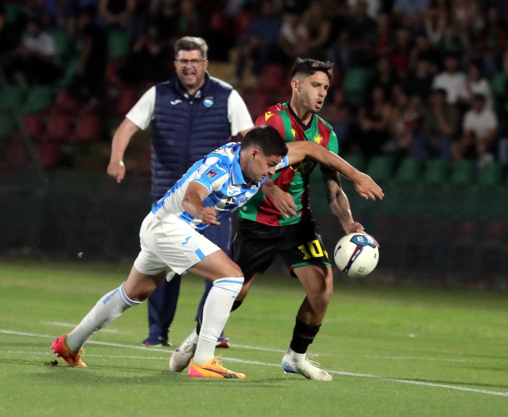 Two soccer players competing for the ball on a green field, with one player in a light blue uniform and the other in a red and green jersey. A coach can be seen in the background, expressing excitement.