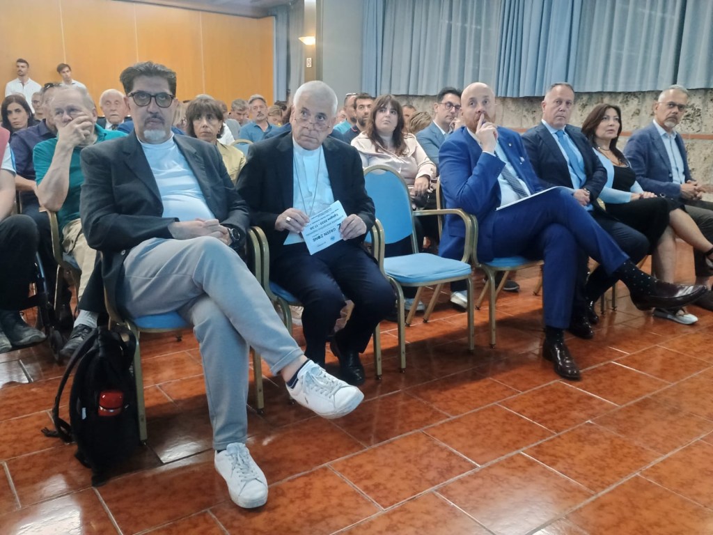 A group of people seated in a conference room, focusing on a speaker. The foreground features three men, with one older man wearing glasses, and others in business attire, while the audience in the background is attentive.