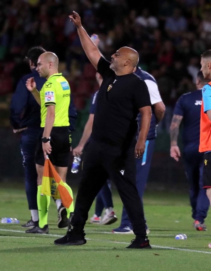 A football coach gestures on the sidelines during a match, wearing a black shirt and pants, with officials and players visible in the background.