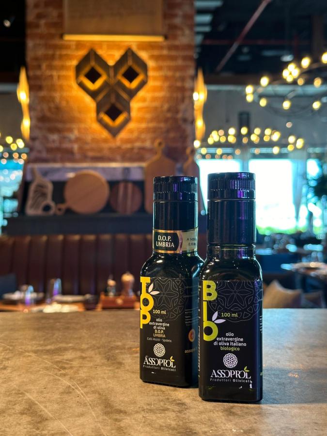 Due bottiglie di olio extravergine di oliva DOP Umbria e biologico, con etichette visibili, posizionate su un tavolo in un ristorante elegante con sfondo arredato.