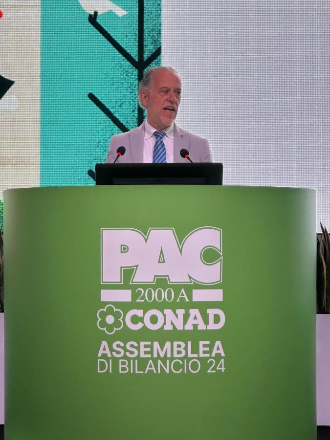 Un relatore parla durante l'Assemblea di Bilancio 2024 di PAC 2000A Conad, con un grande schermo e un pannello verde con logo e testo informativo sullo sfondo.