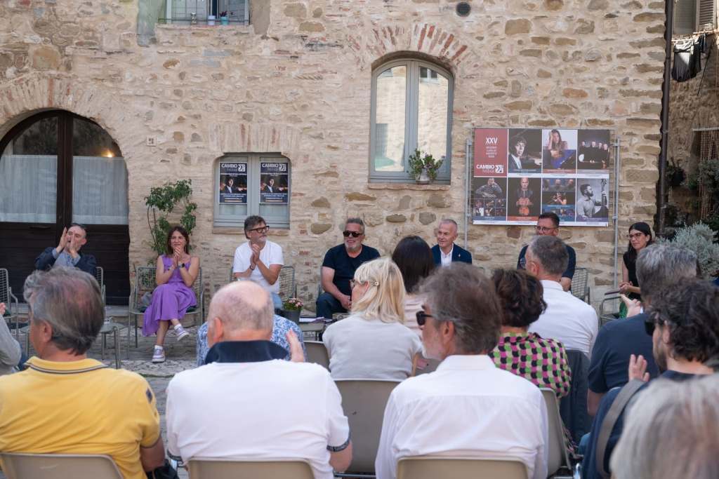 Presentazione del Cambio Festival di Assisi, con un gruppo di persone sedute che applaudono in un contesto all'aperto con pareti di pietra e poster di eventi sullo sfondo.