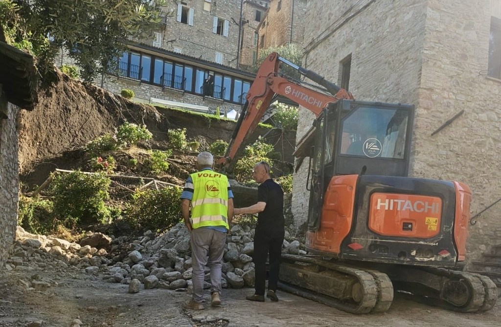 Due uomini osservano un'area di crollo con una macchina escavatrice Hitachi in primo piano. Sullo sfondo, una struttura in pietra con vegetazione circostante.