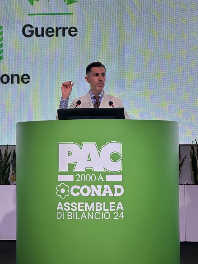 Un oratore al centro di un palcoscenico presenta un discorso durante l'Assemblea di Bilancio 2024 di PAC 2000A Conad, con un grande display verde sullo sfondo che indica il nome dell'assemblea.