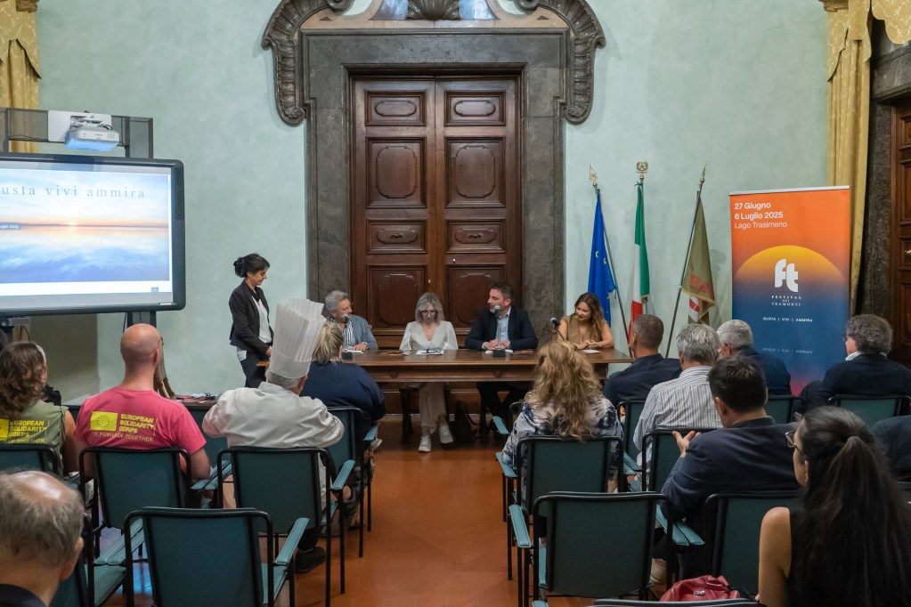 Conferenza stampa per la presentazione del Festival dei Tramonti, con un pubblico attento e relatori al tavolo, in un ambiente elegante con bandiere e un grande schermo sullo sfondo.