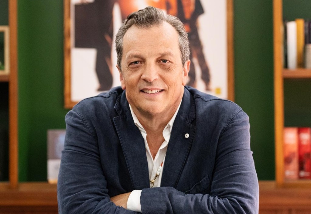 Un uomo sorridente indossa una giacca blu scuro mentre posa con le braccia incrociate su una scrivania. Sullo sfondo si intravedono libri e una decorazione murale.