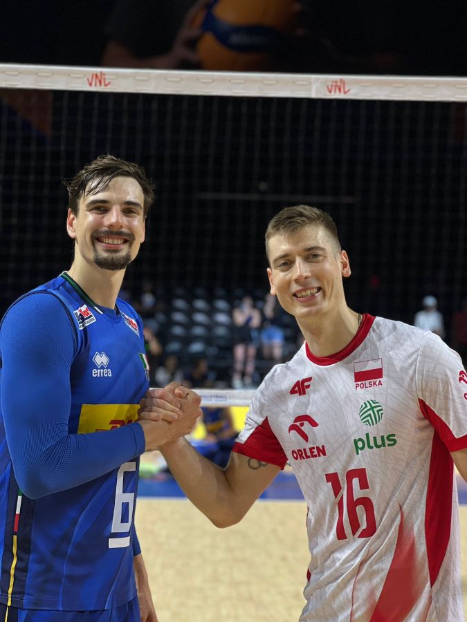 Due giocatori di pallavolo, uno in maglia azzurra e l'altro in maglia bianca e rossa, si stringono la mano sul campo di gioco.