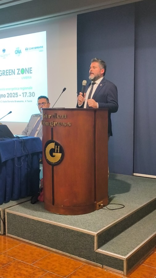 Un relatore in giacca scura parla al microfono durante l'evento 'Green Zone Umbria' al Garden Hotel di Terni, con un altro partecipante visibile sullo sfondo.