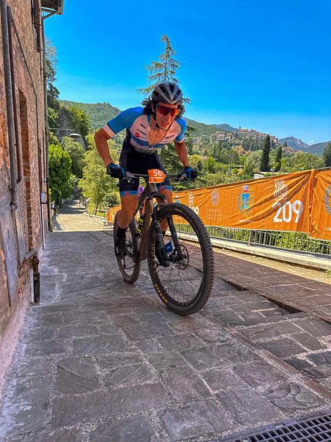 Un ciclista in mountain bike affronta una salita pavimentata durante la 209 Marathon ad Arrone, con un banner dell'evento sullo sfondo e un paesaggio collinare.