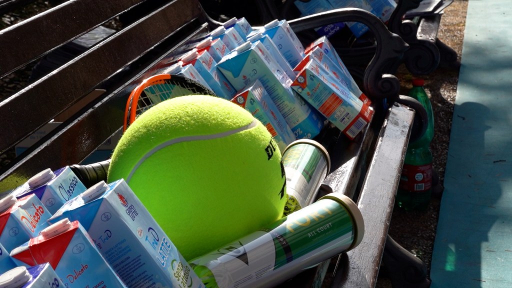 Una panchina con scatole di latte e una pallina da tennis verde in primo piano, mentre in background si intravedono racchette e bottiglie.