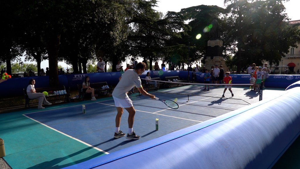 Un giocatore di tennis in azione durante un evento di minitennis ai Giardini Carducci, con bambini e adulti presenti sullo sfondo.
