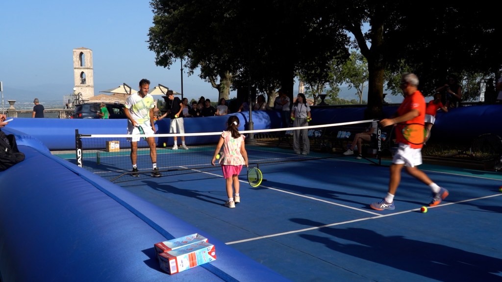 Un momento di minitennis ai Giardini Carducci, con un adulto e una bambina che si scambiano colpi mentre altri partecipanti e spettatori osservano in background.