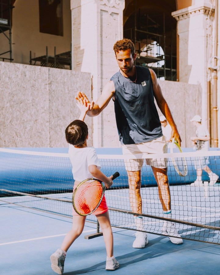 Un giocatore di tennis adulto scambia un high-five con un bambino sul campo da tennis, mentre altri bambini si allenano sullo sfondo.