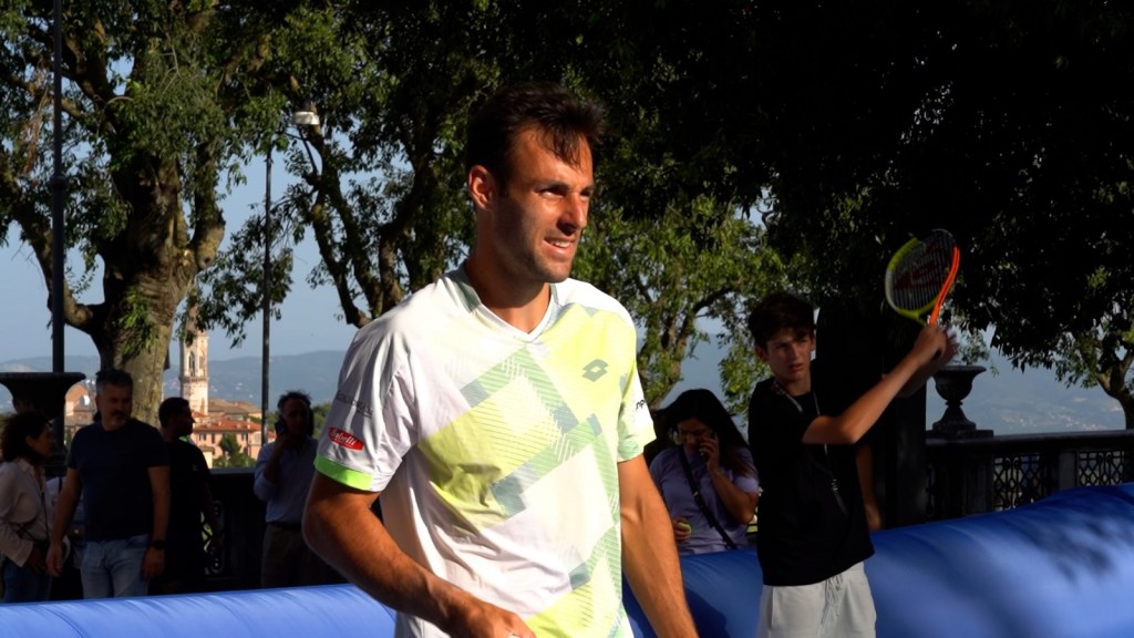 Un giocatore di tennis sorride mentre cammina vicino a un campo, con un giovane che tiene una racchetta sullo sfondo.