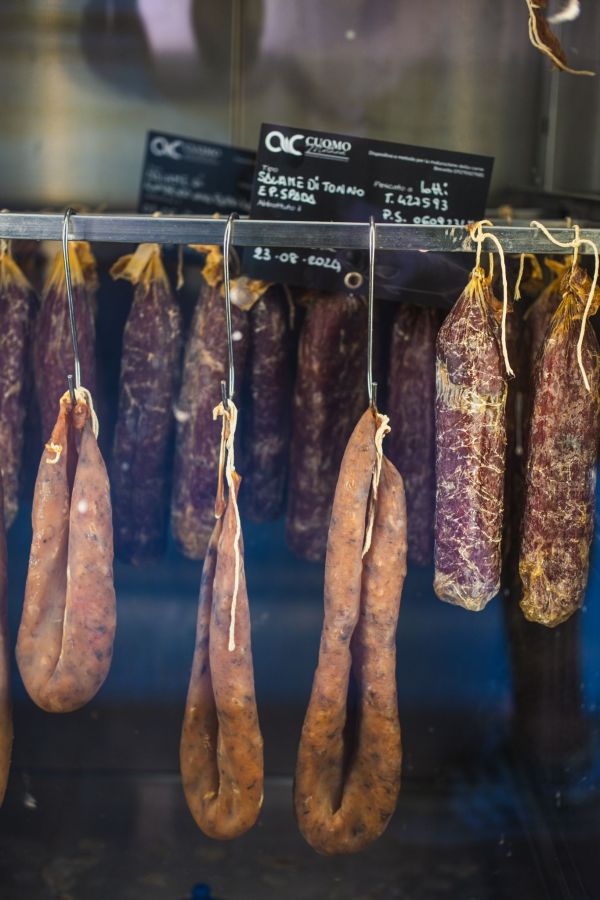 Salumi a base di pesce appesi in un banco refrigerato, con etichette che indicano i prodotti, tra cui salami e salsicce, in un ambiente di pescheria.