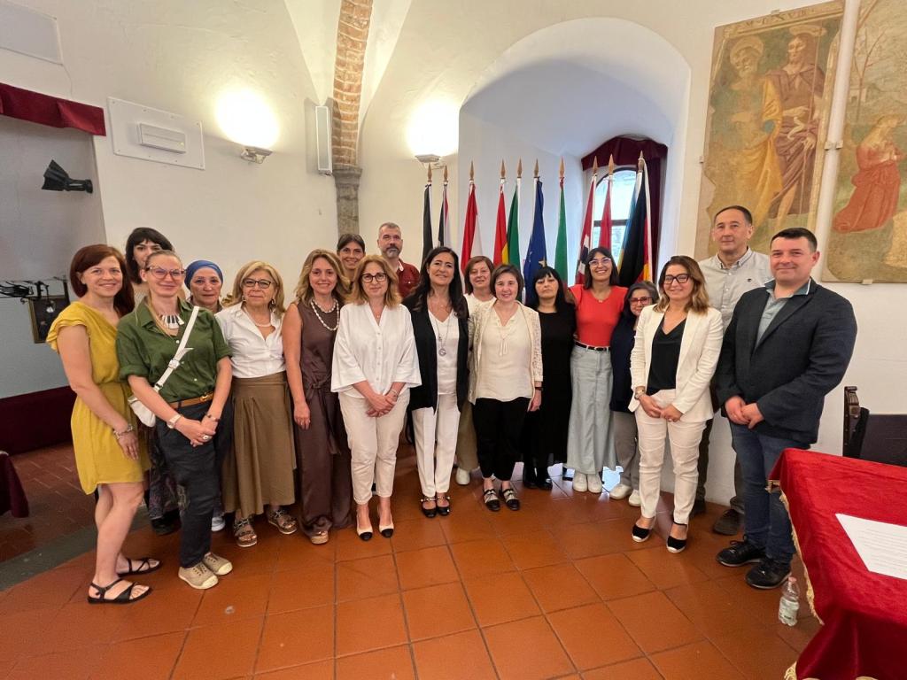 Gruppo di persone in piedi, sorridenti, in un ambiente interno con bandiere sullo sfondo, riuniti per un evento ufficiale.