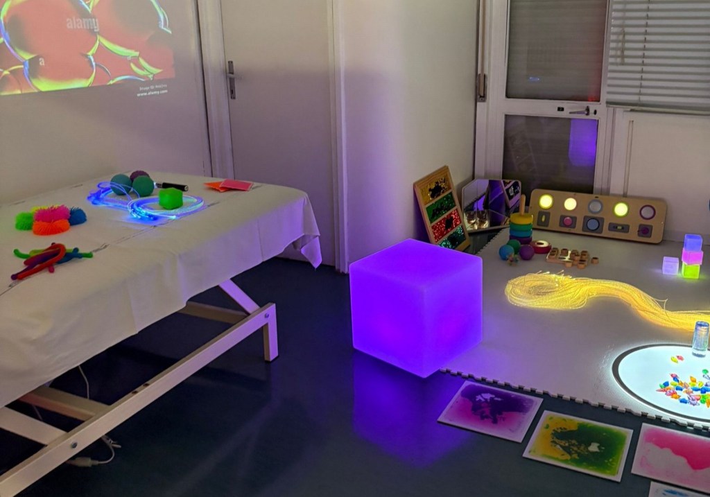 Interno di una stanza multisensoriale con diversi strumenti e materiali colorati, inclusi elementi luminosi e giochi su un tavolo e un pavimento.