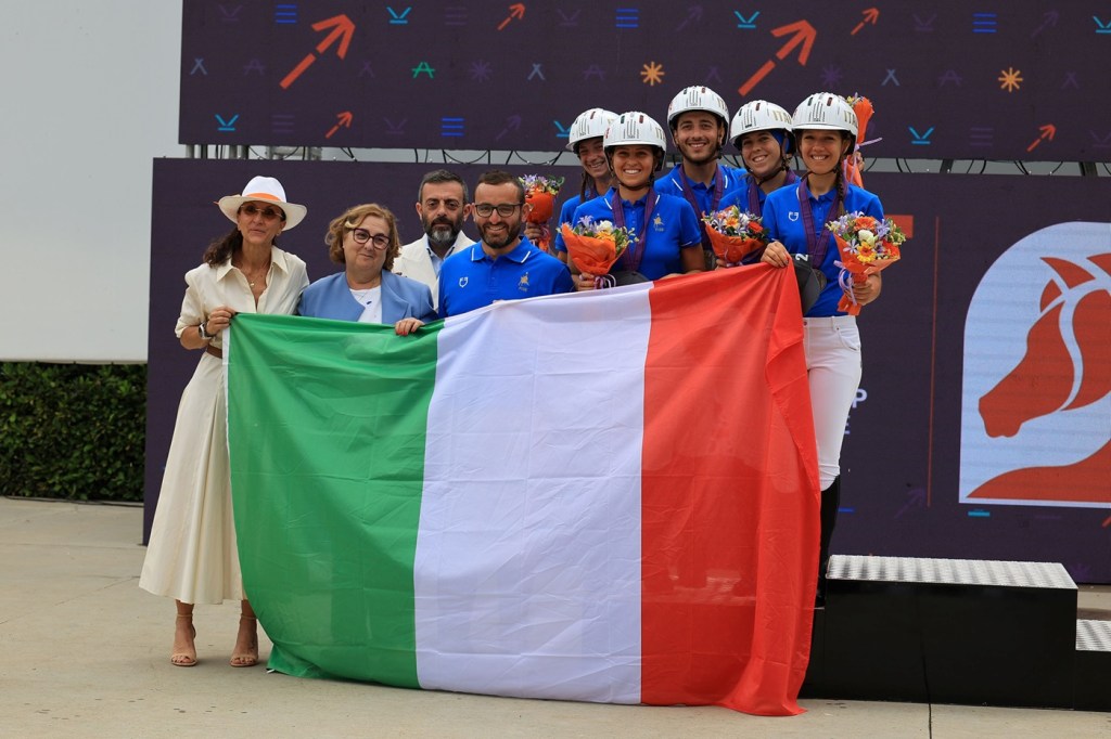 Un gruppo di atleti italiani sorridenti, indossando magliette blu, tengono in mano fiori colorati e una grande bandiera italiana durante una cerimonia di premiazione.