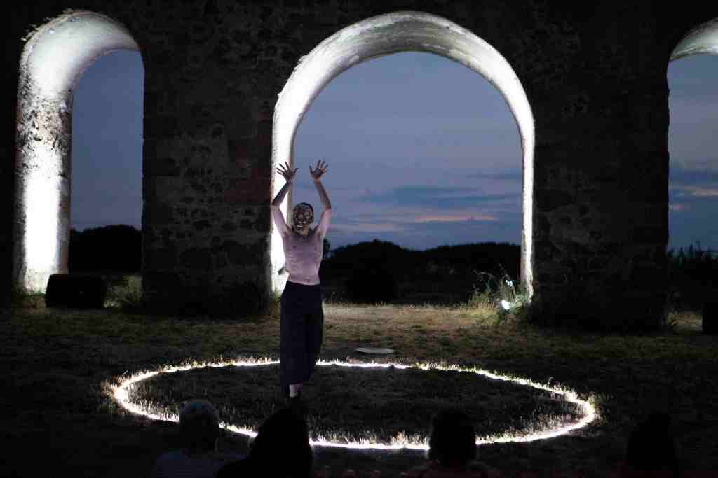 Una performer danza in un ambiente all'aperto al crepuscolo, circondata da archi in pietra illuminati. Il pavimento presenta un cerchio luminoso mentre gli spettatori osservano in silenzio.