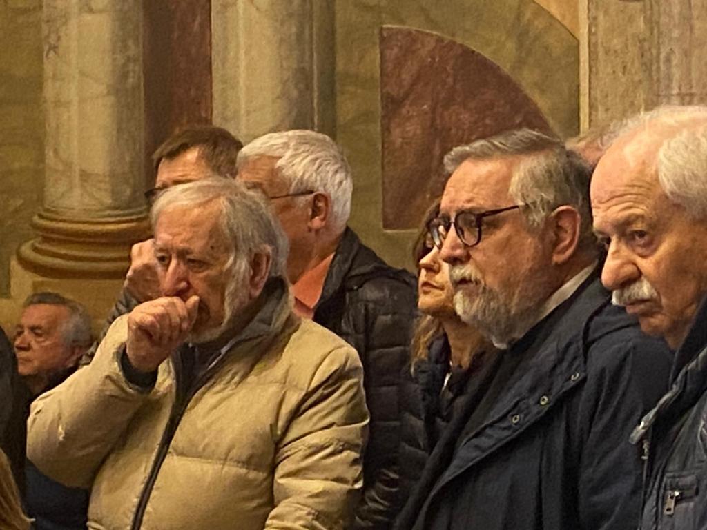 Gruppo di persone anziane in un evento pubblico, alcune con espressioni serie e di riflessione.