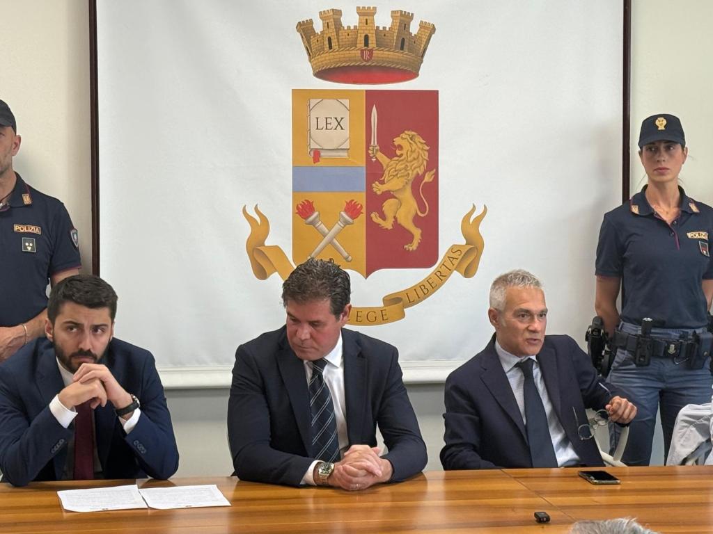 A press conference scene with three men seated at a table, in front of a backdrop featuring the emblem of the police. Two police officers are visible in the background.