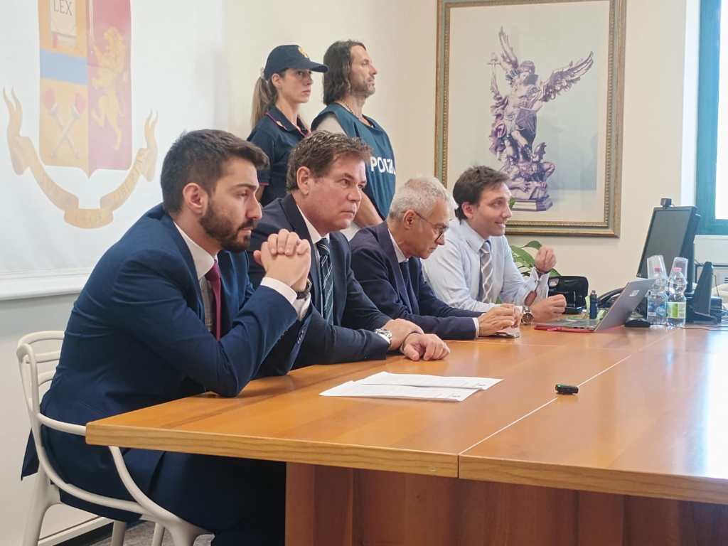 A group of four men seated at a table in a formal setting, discussing matters, with a woman in the background wearing a police vest. A logo with a lion and crossed tools is displayed on the wall behind them.