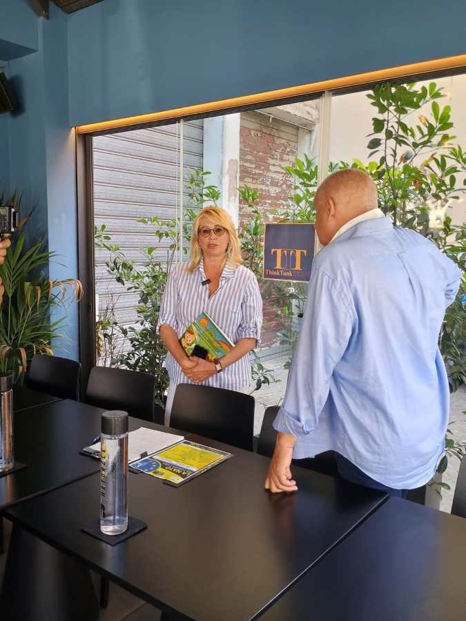 Una donna bionda con occhiali e una camicia a righe interagisce con un uomo, all'interno di un ambiente luminoso, con piante e un logo di ThinkTank visibile sullo sfondo.