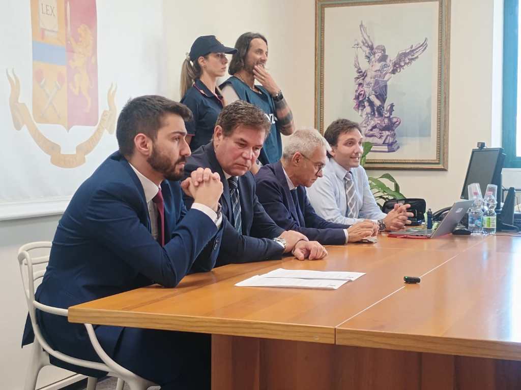 A group of four men seated at a table during a meeting, with a woman standing in the background. The setting includes a wall with a coat of arms and a framed artwork.