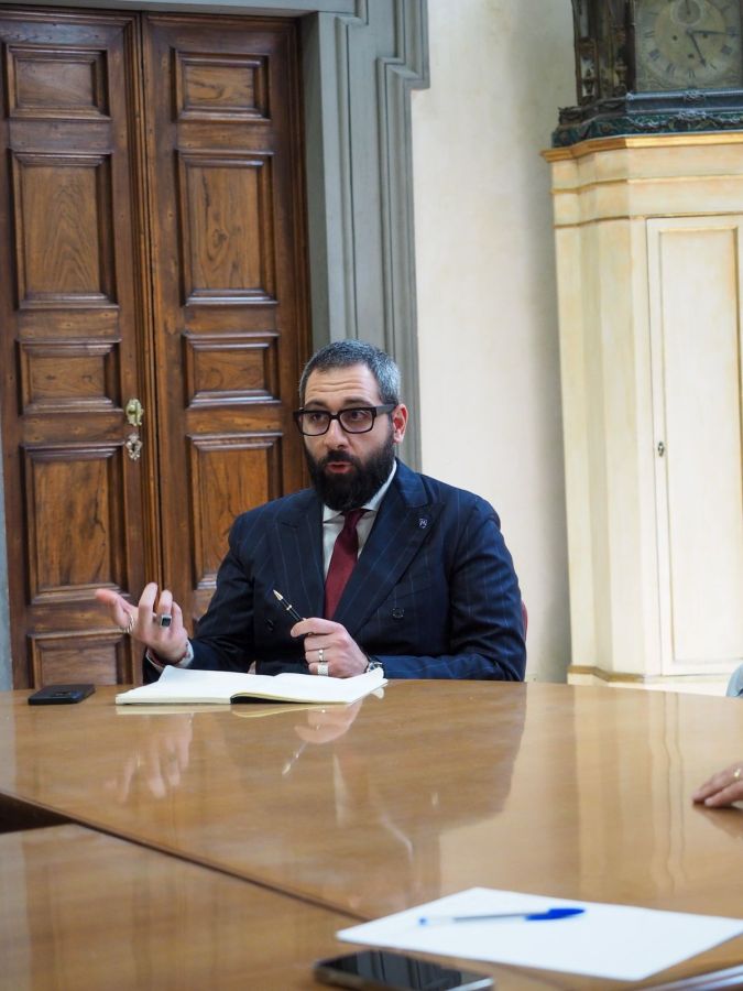 Un uomo in giacca e cravatta discute durante una riunione, mentre tiene una penna in mano. Sul tavolo ci sono un bloc-notes e un dispositivo mobile. Sullo sfondo si vedono delle porte in legno e un mobile di design classico.