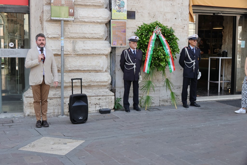 Un rappresentante parla al microfono durante le celebrazioni per l'81° Anniversario della Liberazione di Terni, con due membri delle forze dell'ordine in uniforme accanto a una corona d'alloro decorata con nastri tricolori.
