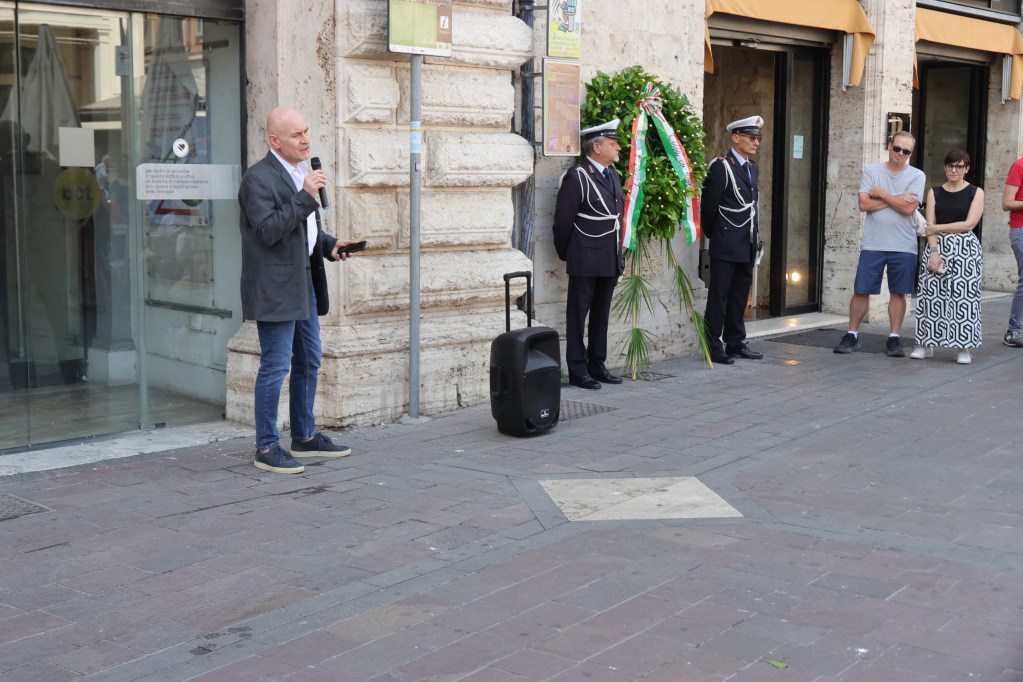 Un uomo parla al microfono durante le celebrazioni per l'81° Anniversario della Liberazione della Città di Terni, con un gruppo di persone e ufficiali in uniforme presenti al suo fianco.