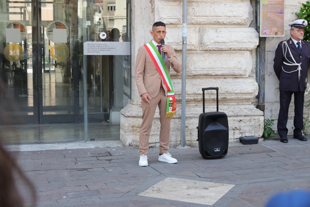 Un uomo con un abito beige e una fascia tricolore parla al microfono durante le celebrazioni per l'81° anniverario della Liberazione a Terni, con alcuni partecipanti e un ufficiale di polizia in uniforme sullo sfondo.