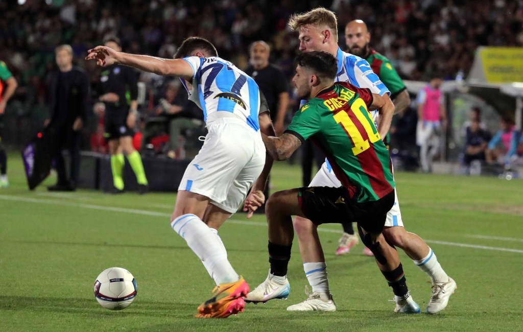 A soccer match scene showing players competing for the ball on the field, with one player in a red and green jersey attempting to regain possession from two opposing players.