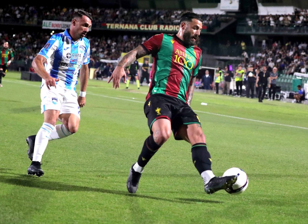 A football match scene featuring two players in action. One player in a blue jersey dribbles the ball while the other, wearing a green and red jersey, attempts to gain possession on a grassy field.