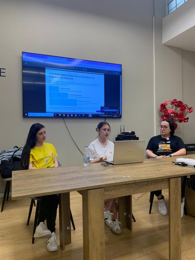 Tre relatrici durante un convegno informativo sull'endometriosi, sedute dietro un tavolo con un laptop aperto. Uno schermo alle spalle mostra un contenuto progettato per la presentazione, mentre un mazzo di fiori è visibile in primo piano.