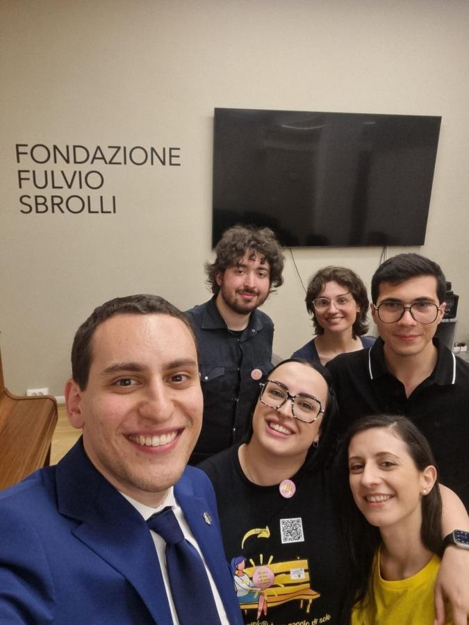 Gruppo di persone sorridenti in un selfie, con un muro sullo sfondo che riporta il nome 'Fondazione Fulvio Sbrolli'.