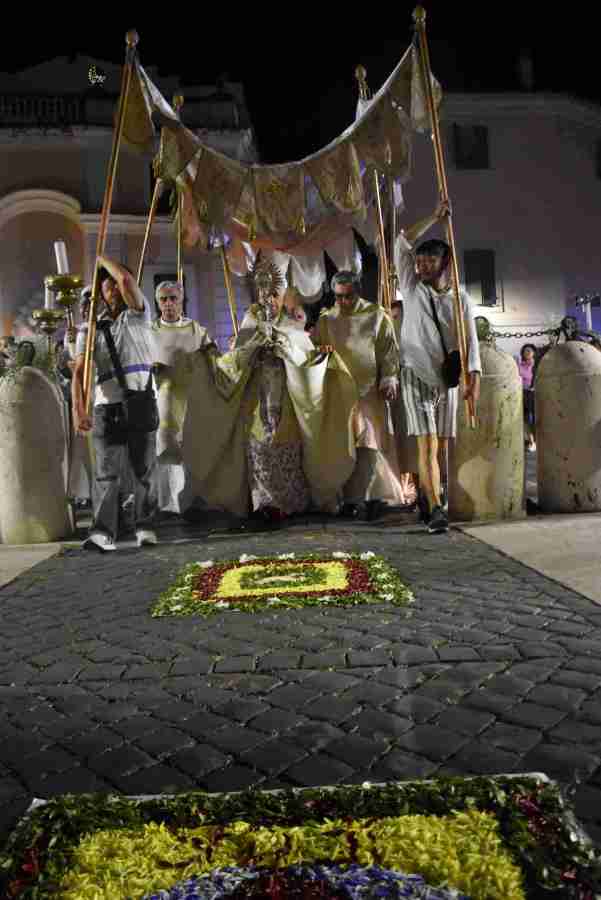 Una processione notturna del Corpus Domini con partecipanti che portano un grande drappo. In primo piano, decorazioni floreali sul pavimento. L'atmosfera è festosa e solenne.