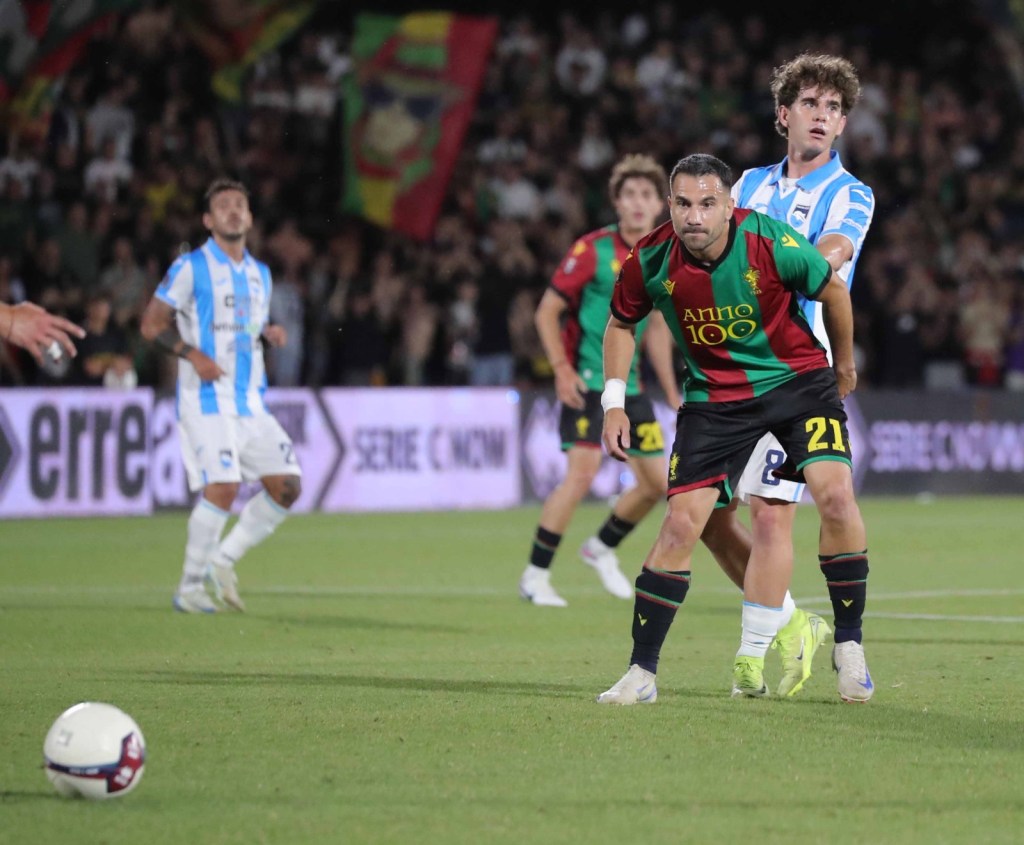 Two football players are engaged in a competitive moment on the field, one in a multicolored jersey and another in a blue and white striped kit. Fans are visible in the background.