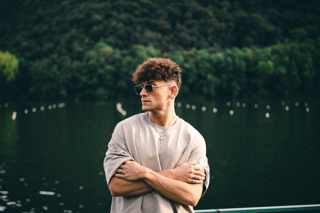 Giovane artista Federico Miotto, conosciuto come Flow Cost, in piedi davanti a un lago circondato da vegetazione, indossa occhiali da sole e una maglietta grigia mentre incrocia le braccia, esprimendo un atteggiamento riflessivo.