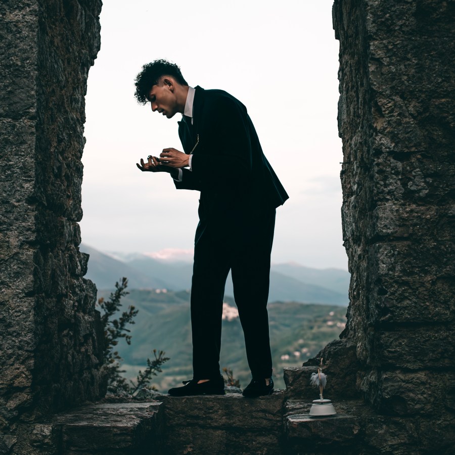 Un giovane uomo vestito elegante si trova in un ambiente d'epoca, all'interno di una struttura mineramente rovinata, mentre guarda un oggetto con espressione pensierosa.